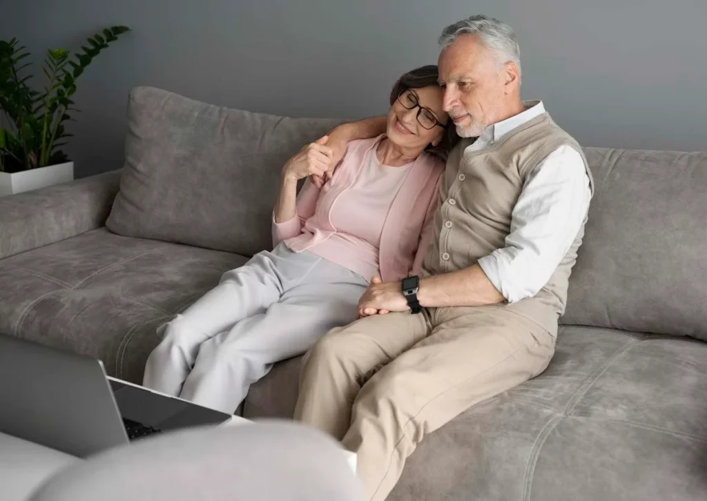 A senior couple sitting together on a comfortable gray sofa, smiling while looking at a laptop in a clean, modern living room